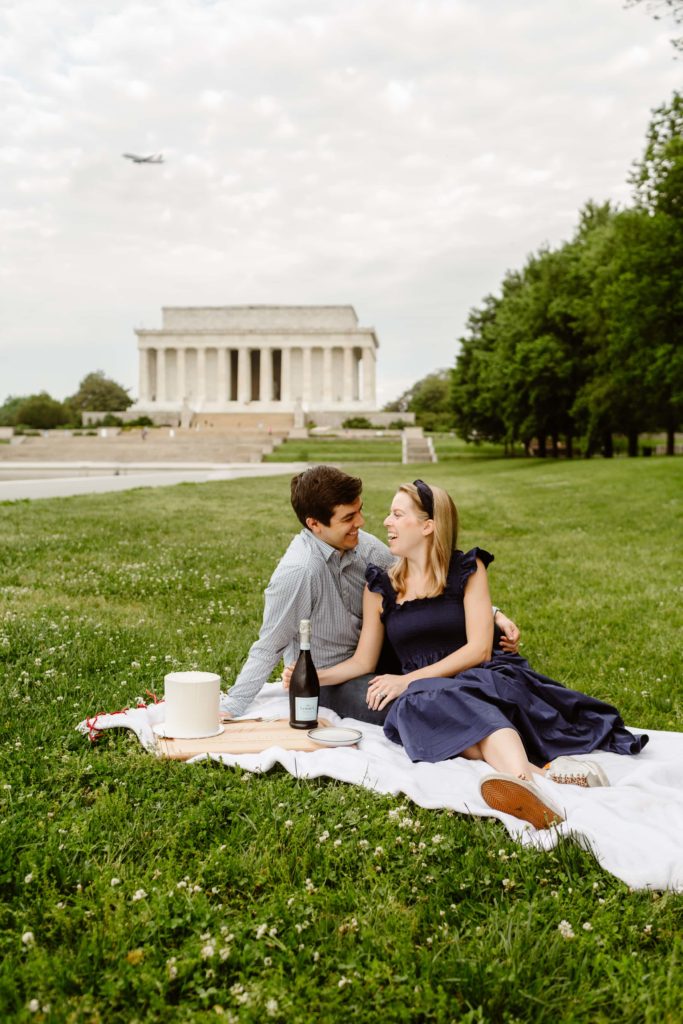 Couple sit on the grass with a romantic picnic, cake, and champagne during a DC engagement photoshoot at the Lincoln Memorial.