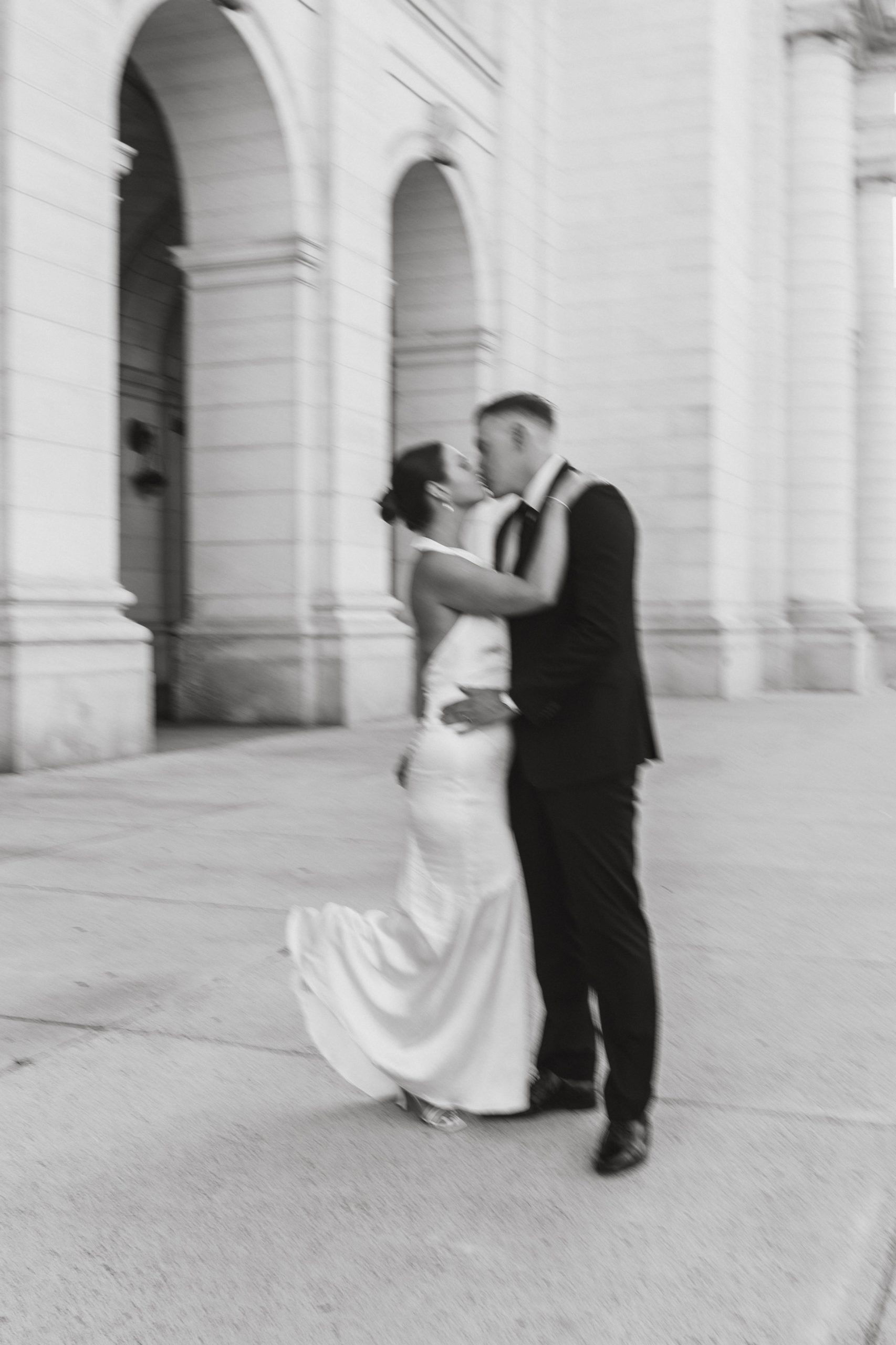 black and white blurry vintage photo of bride and groom kissing in front of white marbled Union Station building with her foot kicked up. 