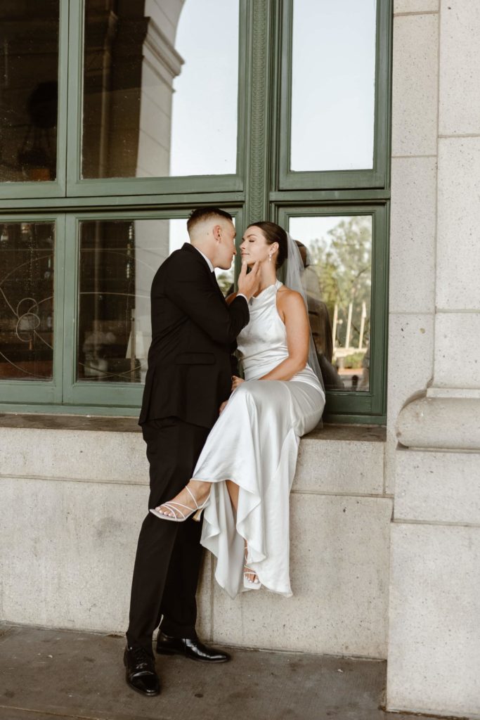 Bride sits on green windowsill at Union Station while groom stands in front of her pulling her in for a kiss.