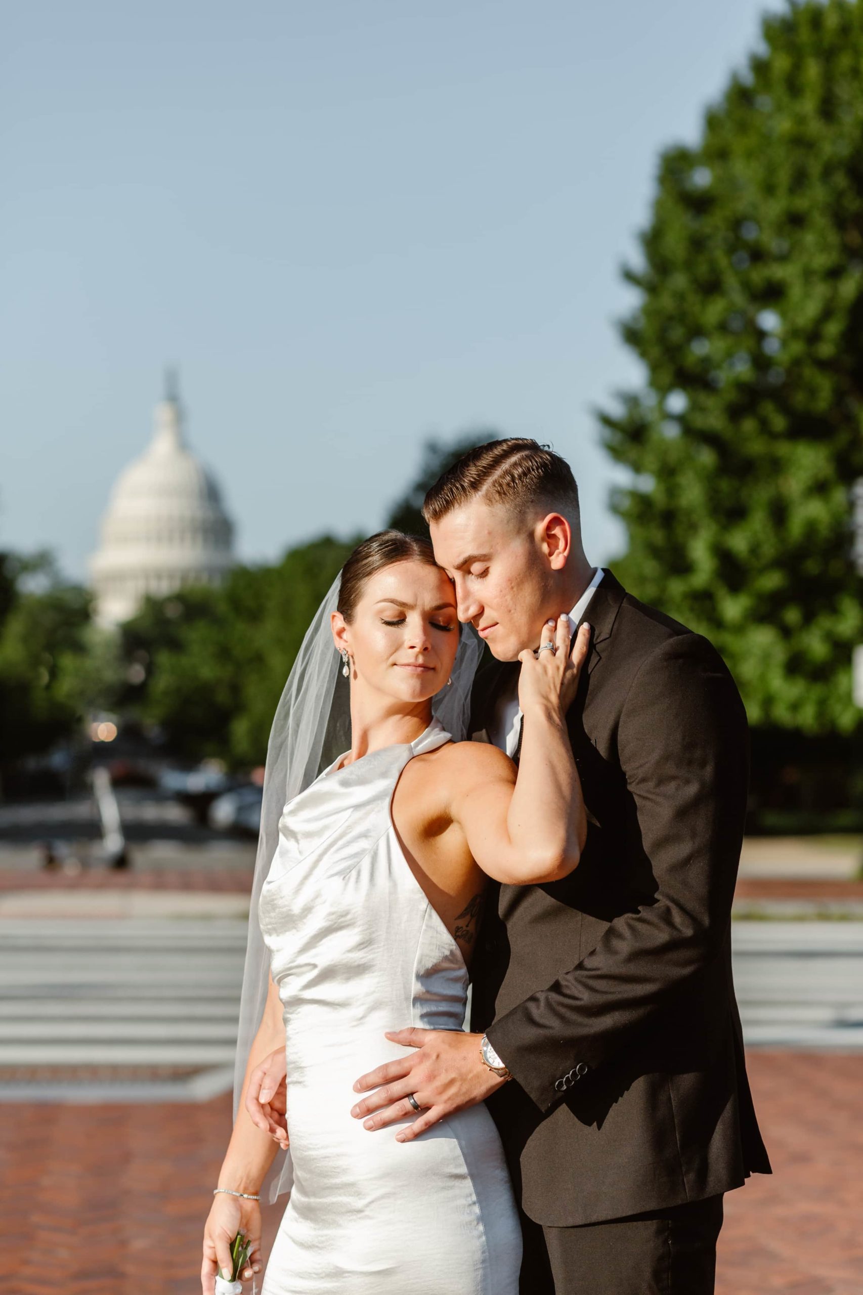 Groom stands behind bride in the sun after their wedding at Union Station with the U.S. Capitol building behind them. 