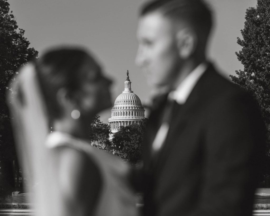 Groom stands behind bride in the sun after their wedding at Union Station with the U.S. Capitol building behind them.