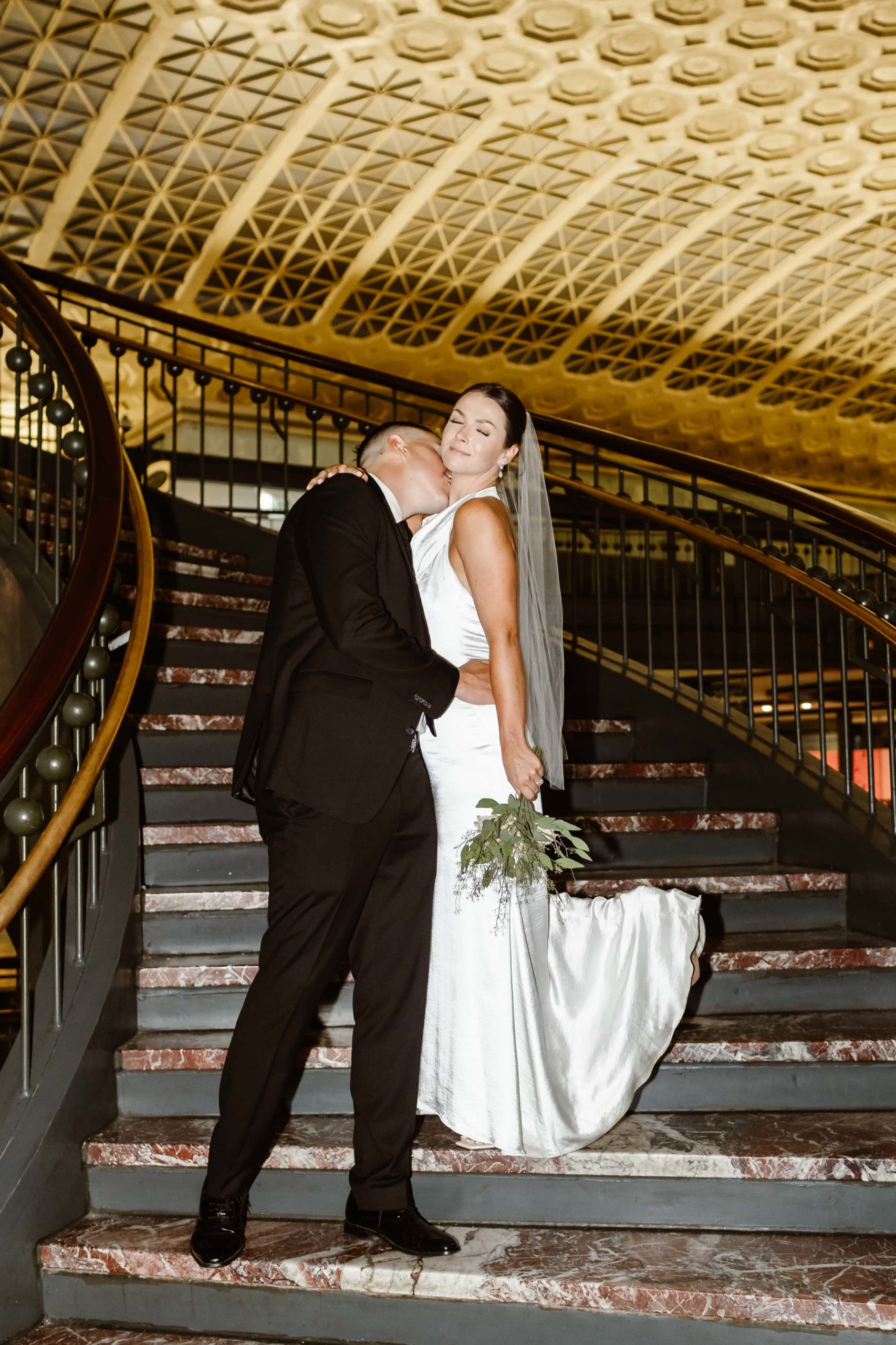 Groom kisses bride's neck while standing on a staircase in Union Station with flash photography