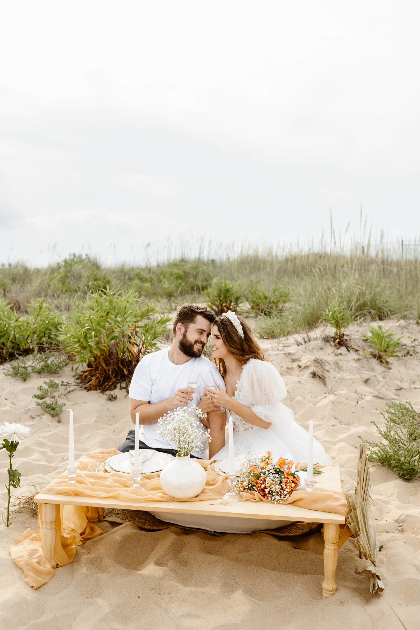 Bride and groom kneeling together on the beach at Bohemian luxury picnic on in Virginia Beach in a short wedding dress while cheering champagne glasses.