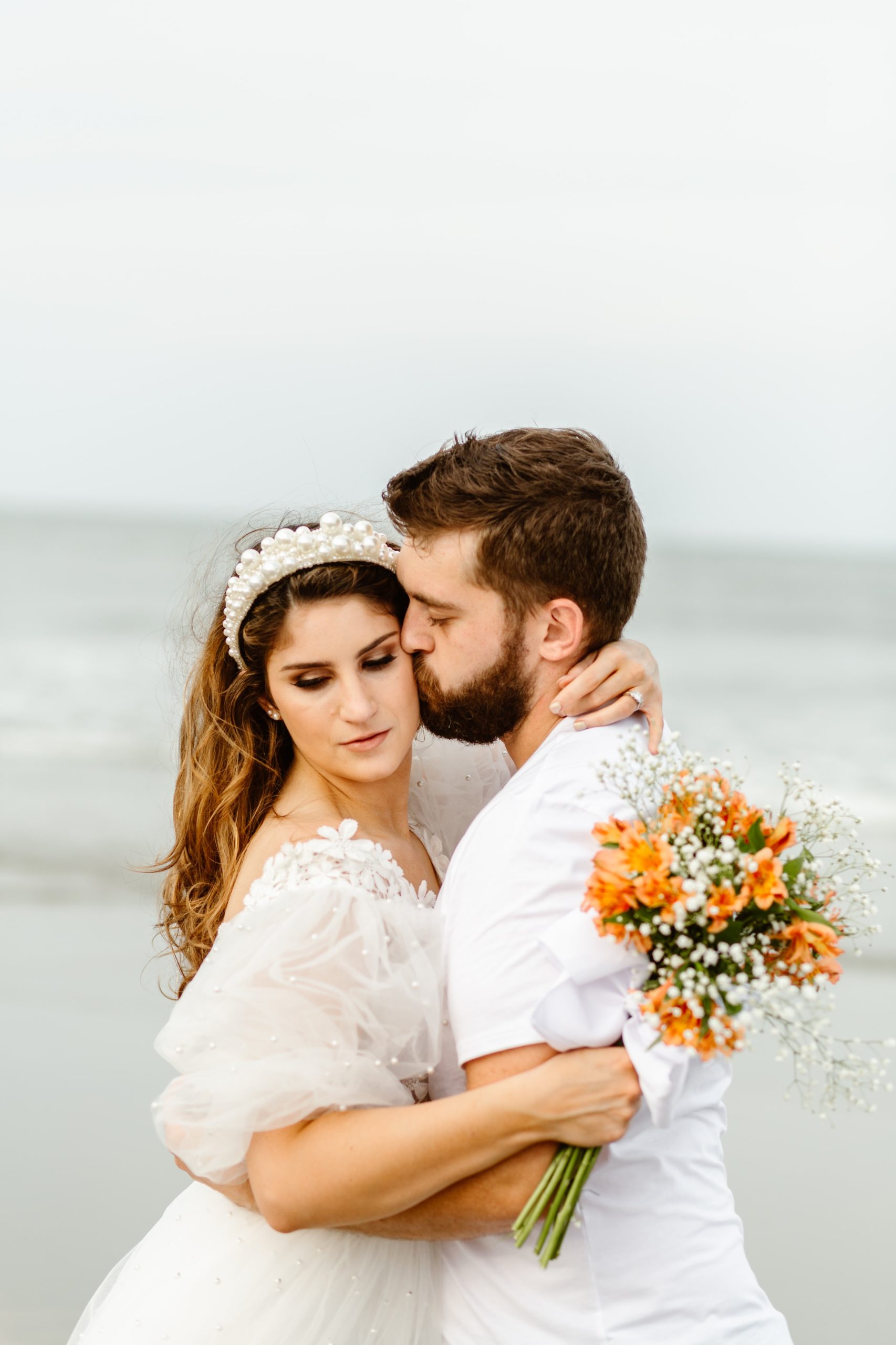 groom kissing bride's cheek while holding orange flower bouquet around his neck on the beach