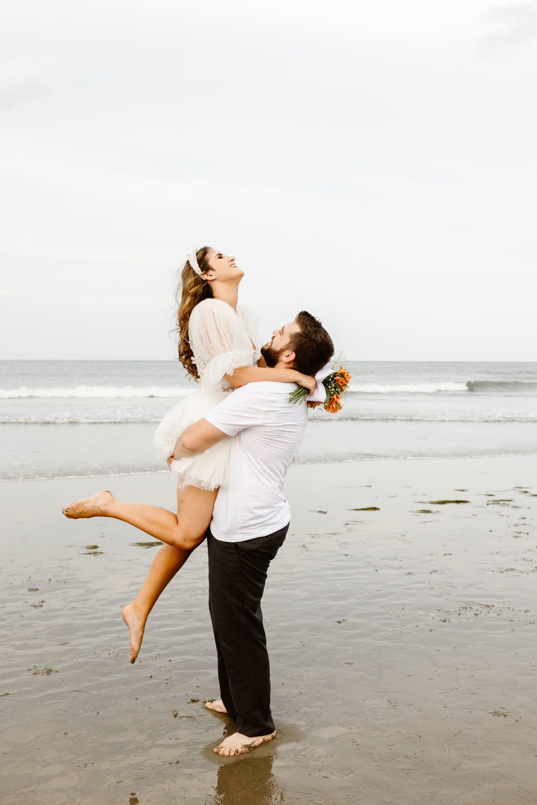 Groom lifting bride in the air as she laughs on the beach in short wedding dress