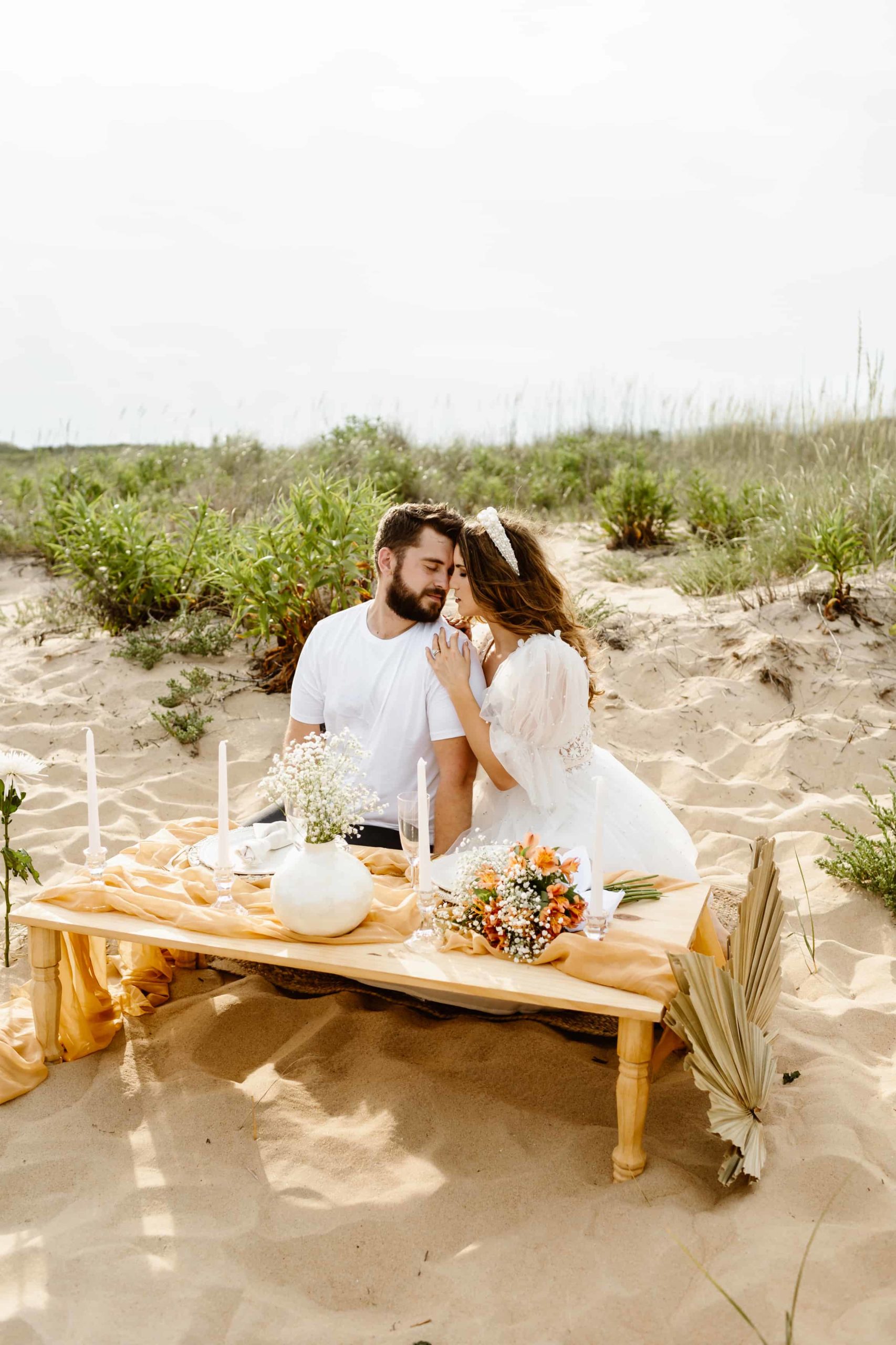 Bride and groom cuddling together on the beach at Bohemian luxury picnic on in Virginia Beach in a short wedding dress.