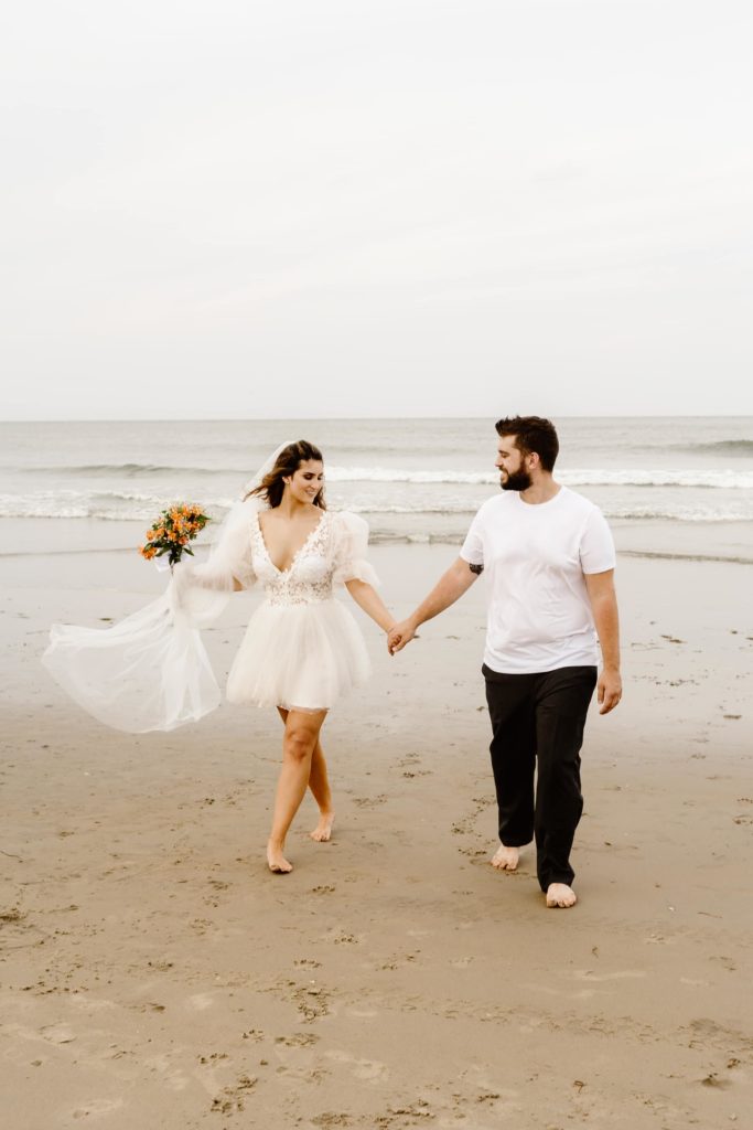 Bride and groom walk in the sand along the beach with bouquet and wedding veil