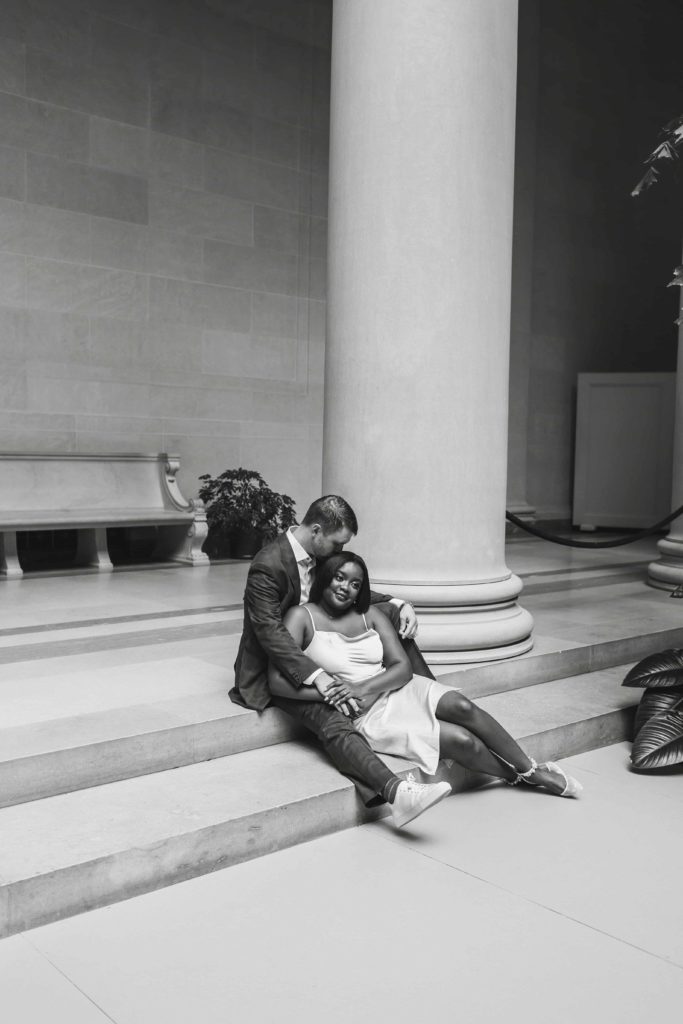 Black and white photo of man and woman sitting together on marble steps in art museum. 