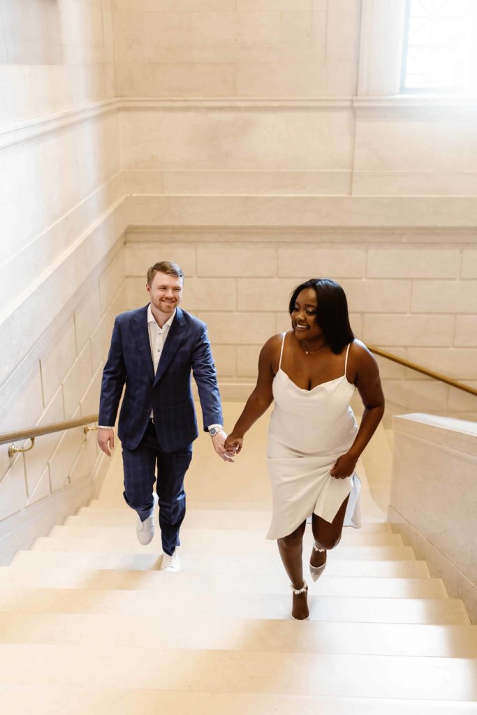 Bride in white stain dress walks up marble staircase leading groom up the stairs in an art museum. 