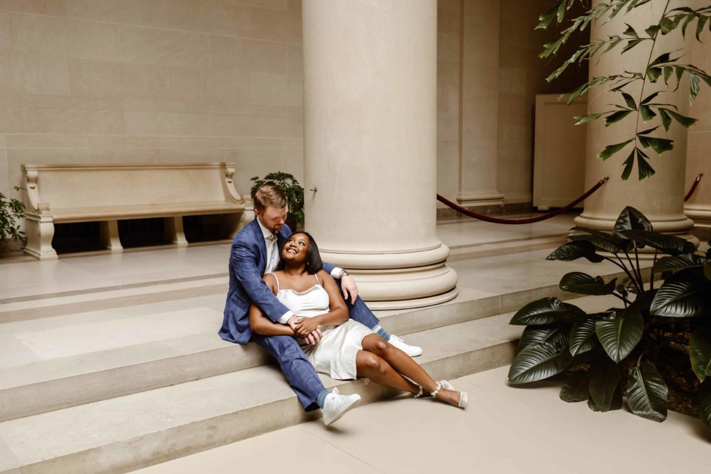 Bride in a satin dress sits on marble stairs leaning against groom who is wearing a blue suit in the National Gallery of Art garden. 
