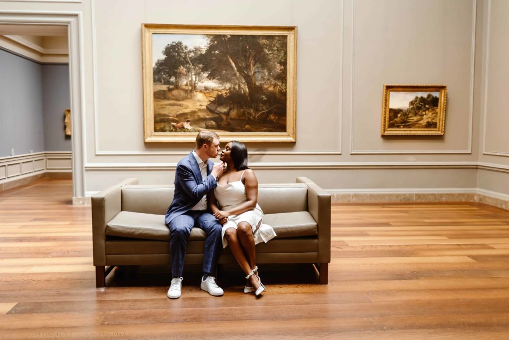 Couple sits on a couch in front a huge painting hind them during their engagement session in the National Gallery of Art in Washington DC. 
