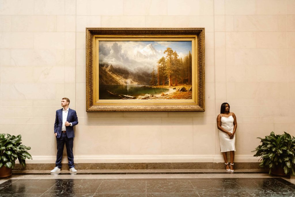 Man and woman stand on opposite ends of a landscape painting during a National Gallery of Art engagement session. 