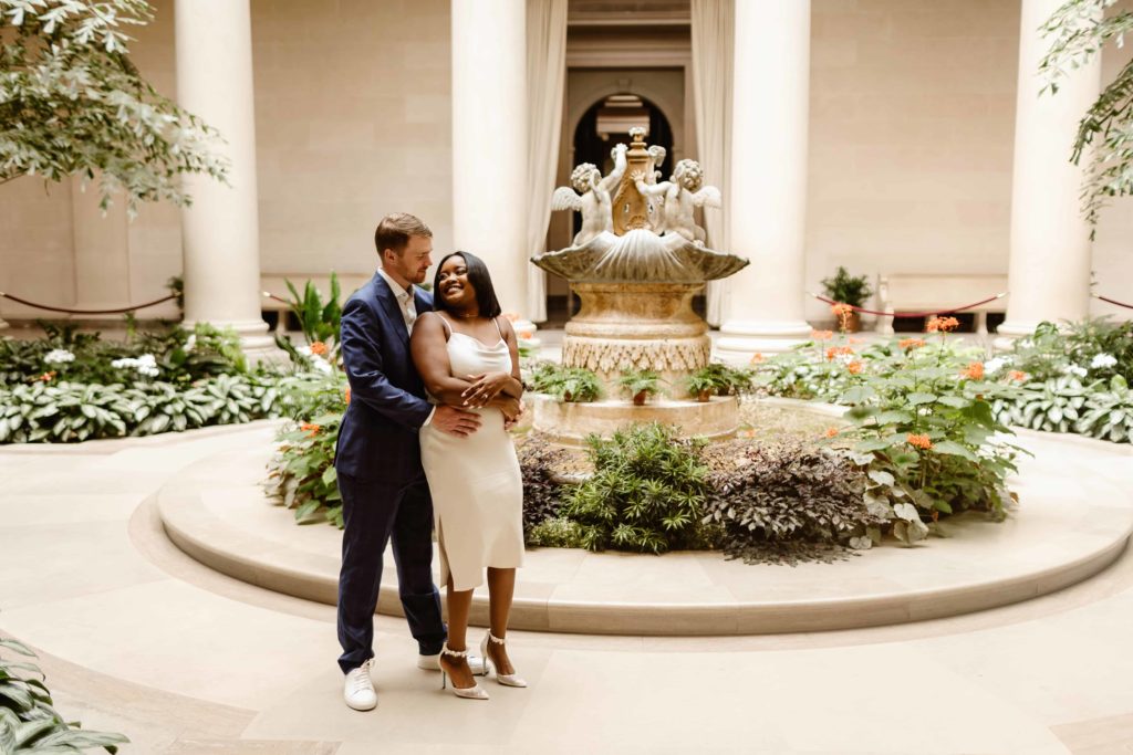 Man and woman embrace in front of a garden fountain in the National Gallery of Art during an engagement session. 