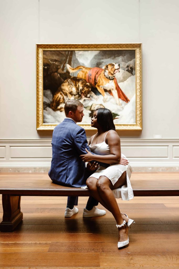 A couple sits on a bench in front a painting of dogs in the National Gallery of Art. 