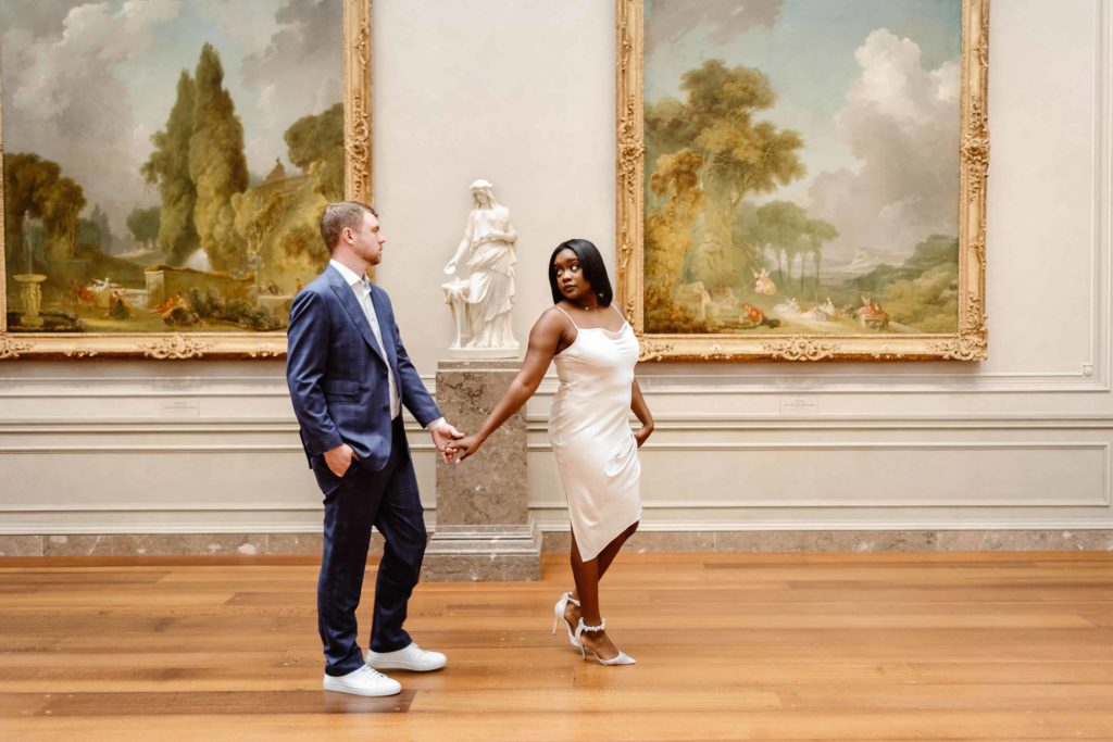 Bride in white satin dress and white heels leads the groom across the room in front of paintings and a sculpture in an art museum in DC.
