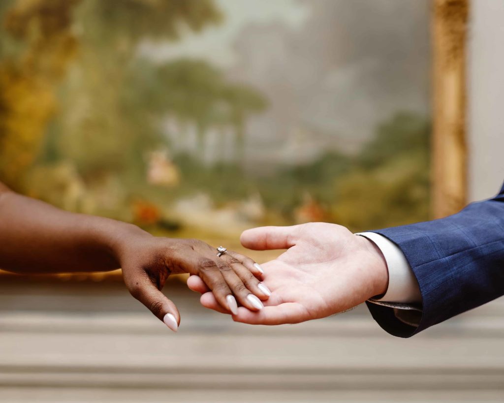 A close up photo of a man and woman's holding hands with engagement ring in an art museum. 