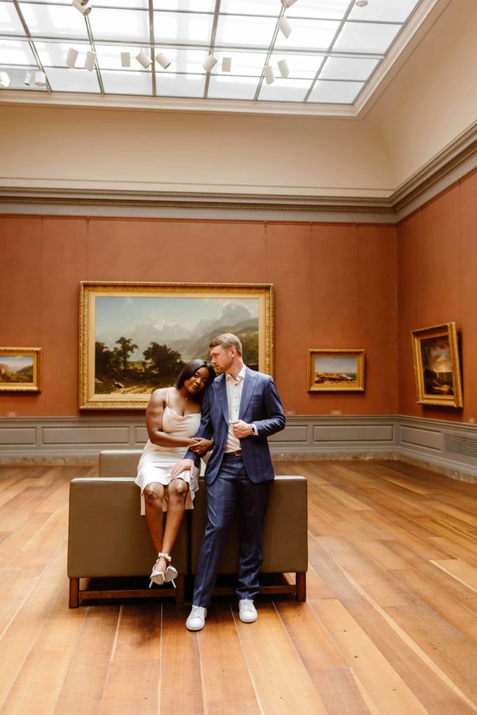 Bride sits on arm of a couch while groom stands next to her as they are in a red painted room of the National Gallery of Art