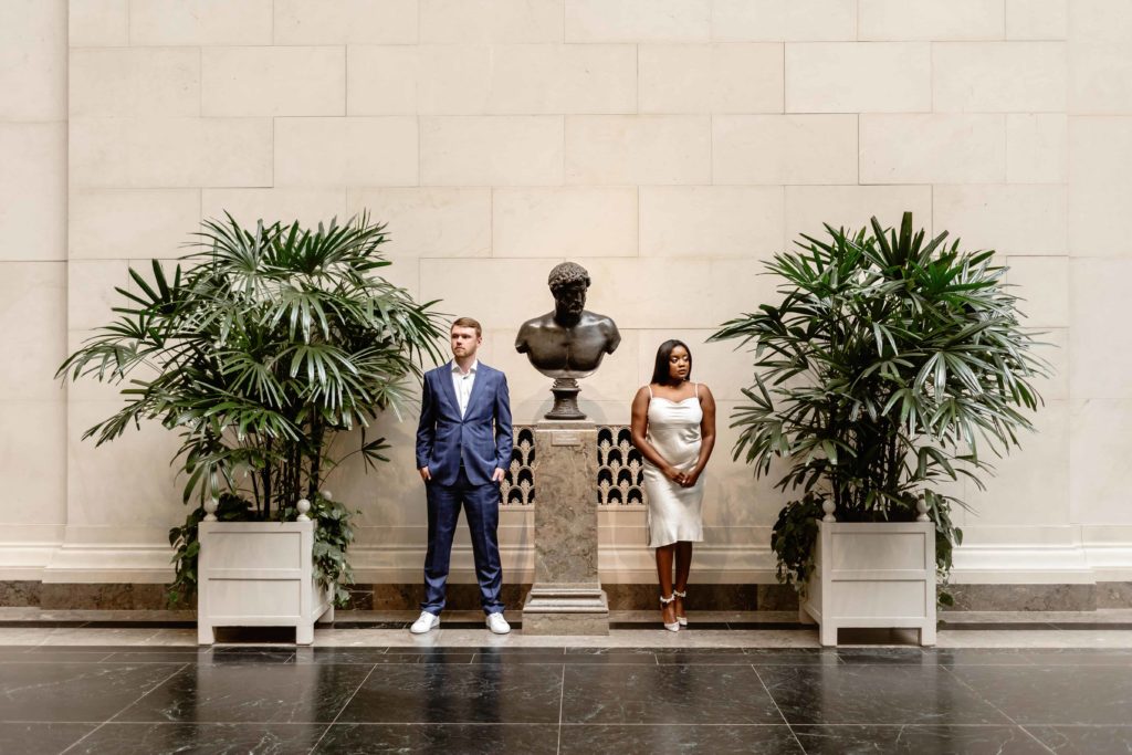 Engaged couple stand on opposite sides of a marble bust statue surrounded by plants in the National Gallery of Art in DC.