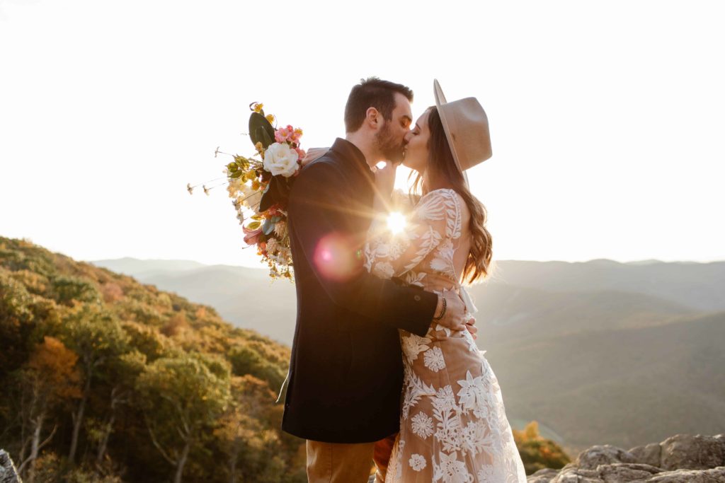 Wedding couple kiss in the mountains with a sun flare coming in between them. The Bride holds her flowers behind the groom during their Raven's Roost elopement in the Virginia Mountains. 