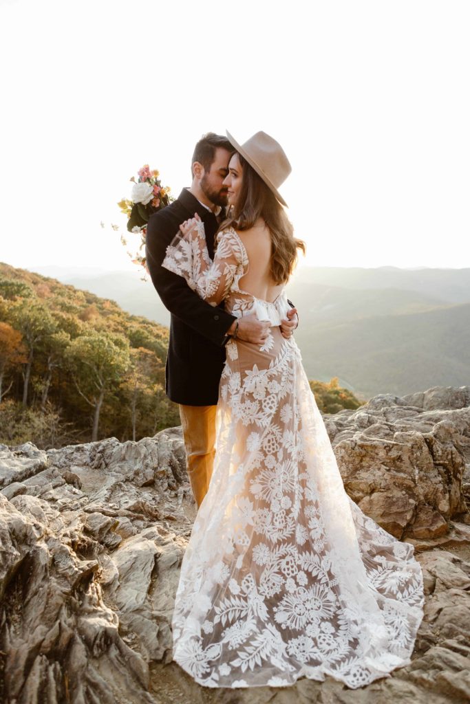 Bride stand with her back to the camera with her bohemian dress fluffed out holding her husband. The wedding couple stands on a cliff in the Virginia mountains during their Raven's Roost Overlook Elopement. 