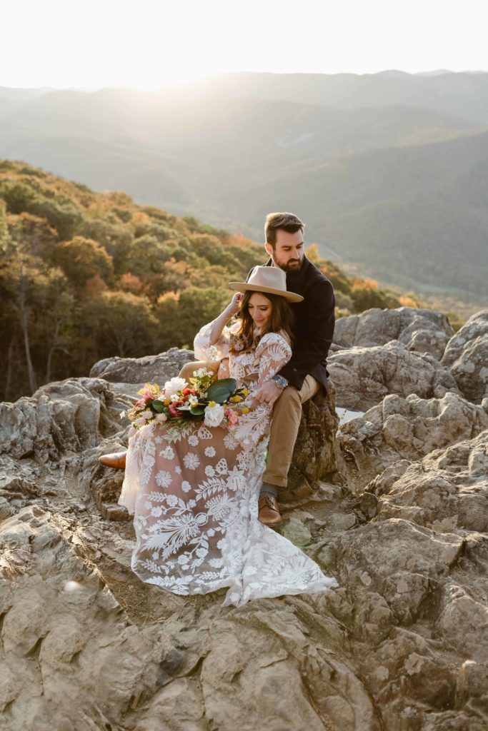 Bride in a boho wedding gown sits on rocks in front of her groom at sunset with the Virginia mountains behind them during their Raven's Roost Overlook elopement. 