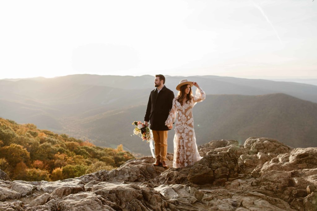 Bride and groom hold hands while looking opposite directions and the bride holds her hat. The wedding couple stand in front of Virginia mountains during their Raven's Roost Overlook Elopement. 