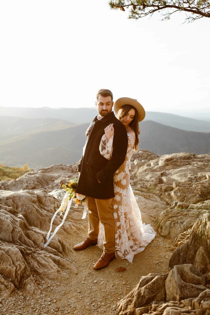 Bride hugs groom from behind while standing in the mountains at sunset during their Raven's Roost Overlook Elopement. 