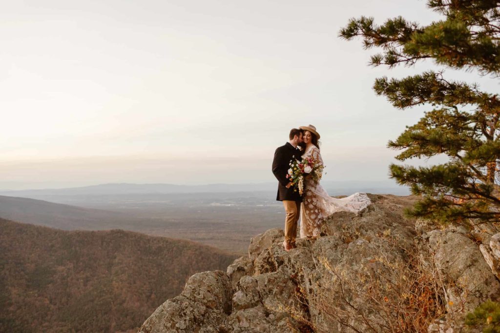 Groom kisses bride's cheek as they stand together holding each other on a mountain cliff in Virginia during their Raven's Roost Overlook Elopement. 