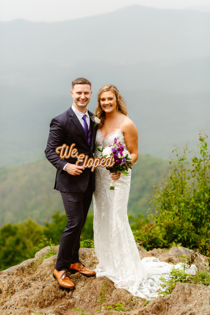 Elopement couple hold a We Eloped sign while standing on a cliff during their Shenandoah National Park Wedding. 