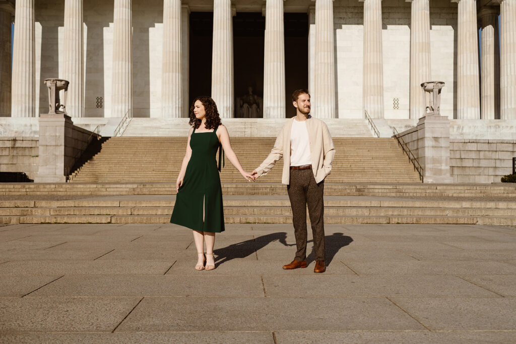 The engaged couple holding hands in front of the Lincoln Memorial as they look in opposite directions