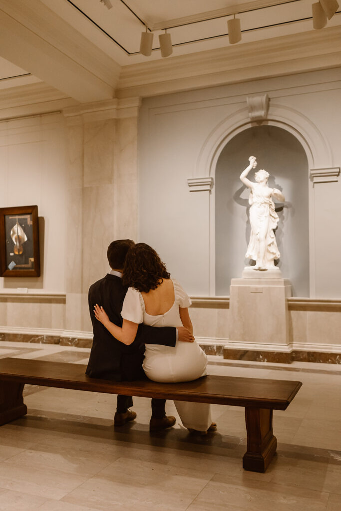 The engaged couple sitting on a bench in the National Gallery of Art looking at a statue together.