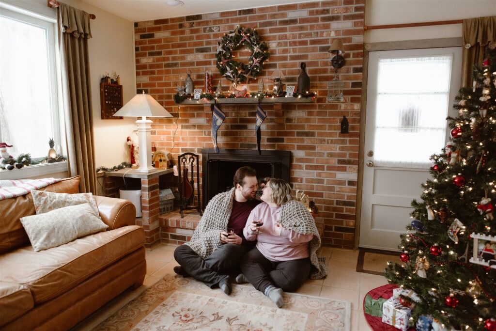 engaged couple snuggled under a blanket next to their fireplace and Christmas tree for winter engagement photos