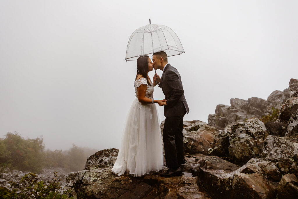 the wedding couple kissing in the rain with their clear umbrella during their rainy wedding day