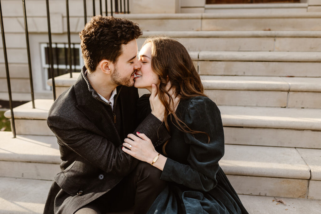 the engaged couple kissing as they sit on the front steps of the Great Marsh Estate