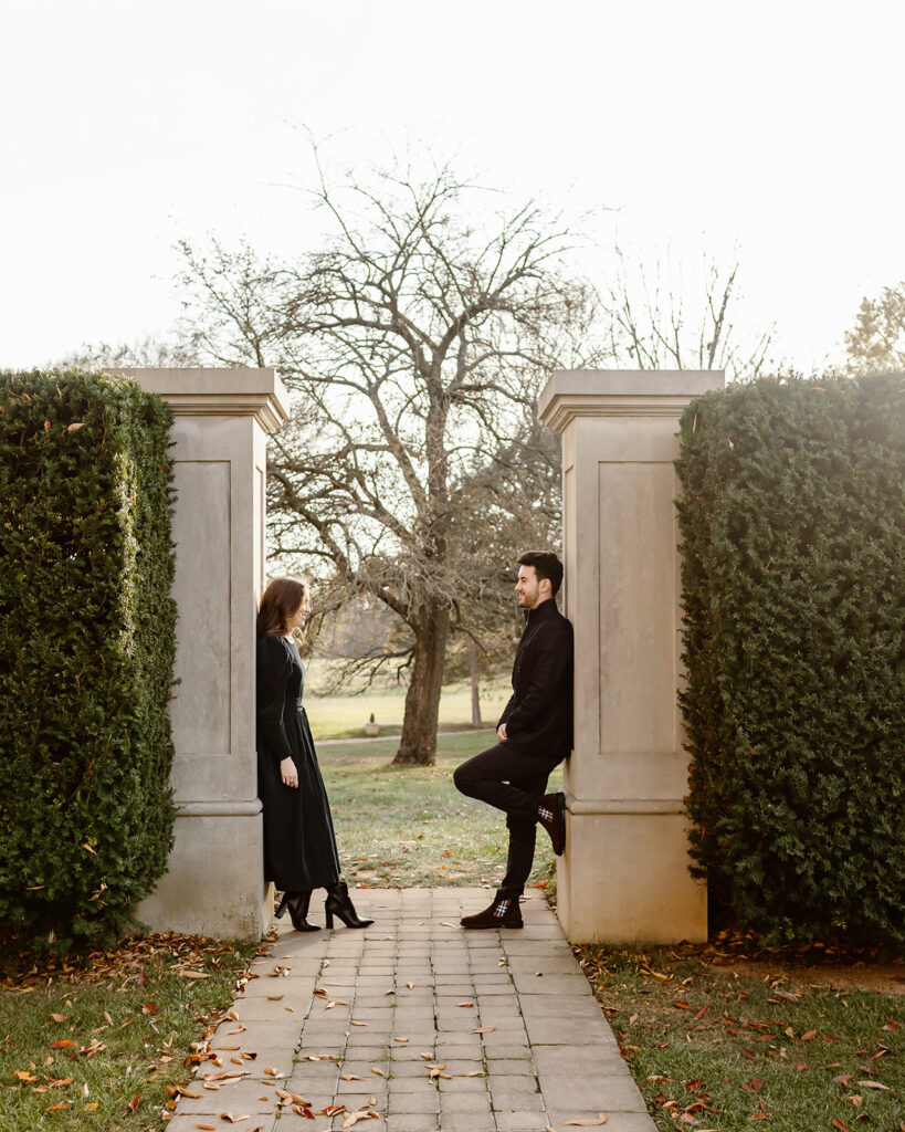 The engaged couple leaning against the pillars in the garden