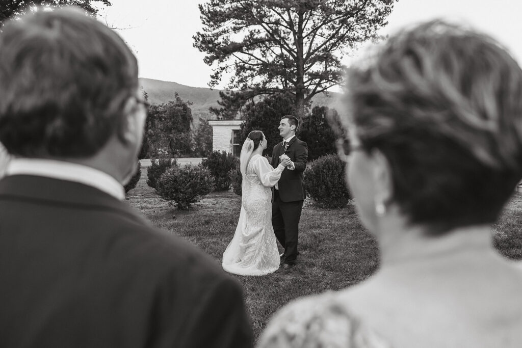 Black and white photo of wedding couple during their first dance and the photo is taken between the bride's parents.