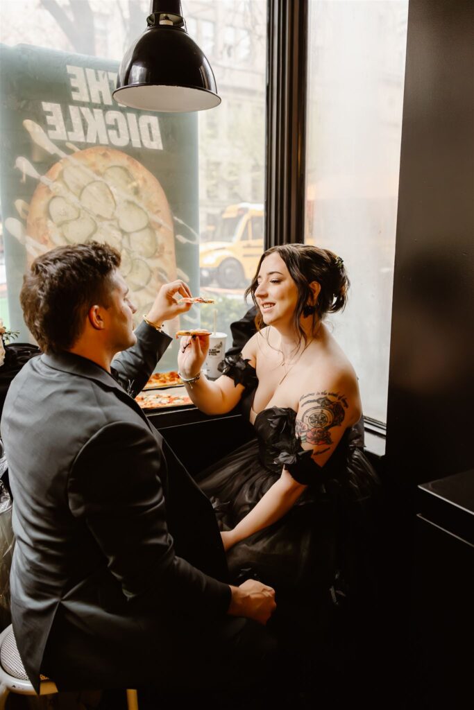 bride and groom in gothic elopement attire sharing a pizza in a cozy cafe