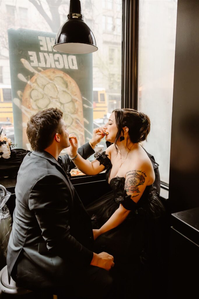 bride and groom in gothic elopement attire sharing a pizza in a cozy cafe