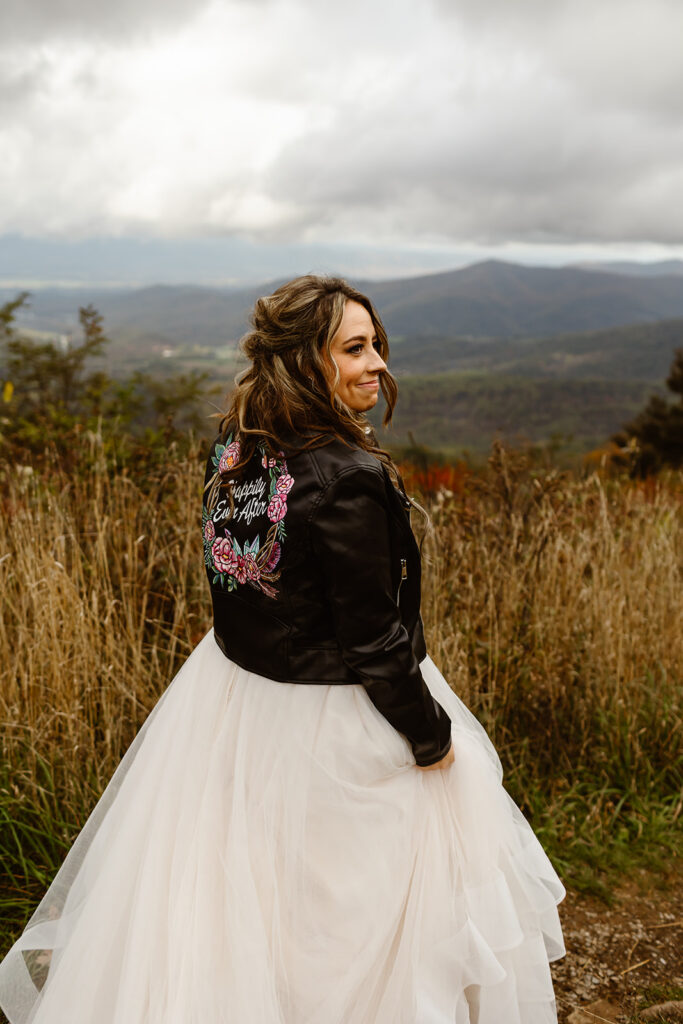 smiling bride in a lush field with a custom leather jacket saying "happily ever after"