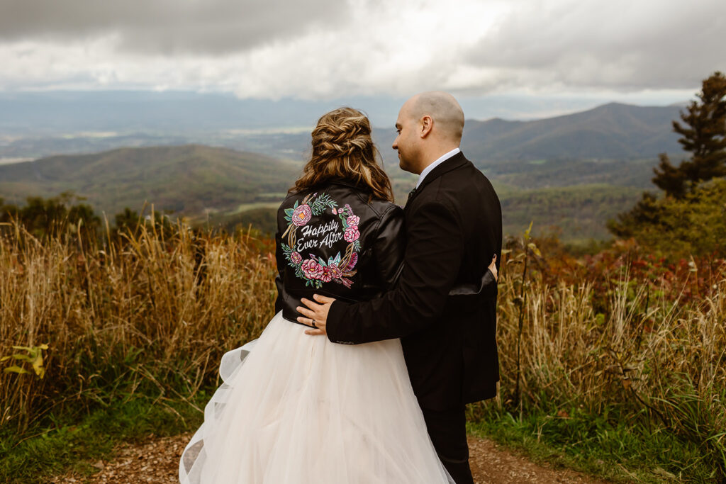 smiling bride and groom in a lush field with bride wearing a custom leather jacket saying "happily ever after"