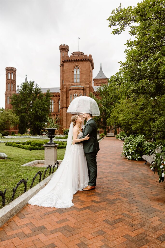 sweet bride and groom under clear umbrellas during rainy moments at the smithsonian castle garden