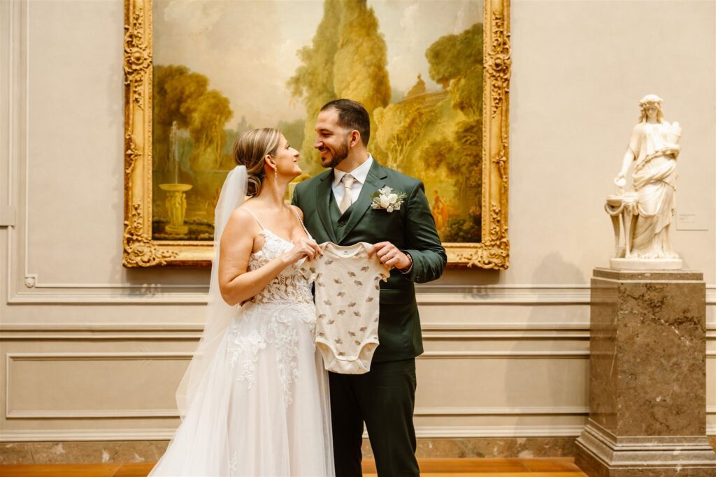 bride and groom holding a tiny baby onesie in front of a painting at the national gallery of art